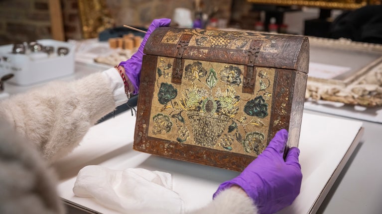 A metal bound round topped casket is being held in gloved hands over a desk. It is decorated with colourful pattern of flowers and leaves